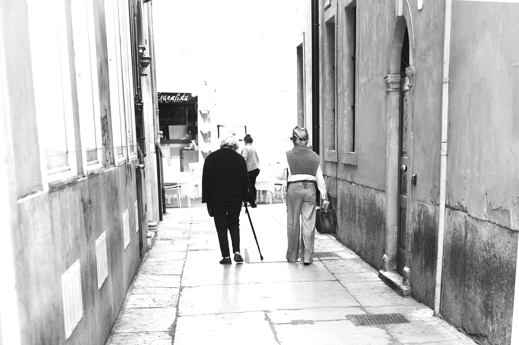 Eine alte Person mit Stock geht eine enge, helle Gasse hinunter, von hinten gesehen