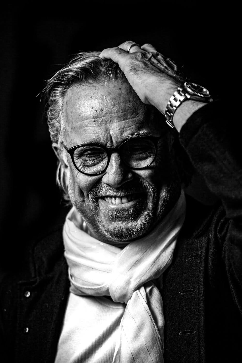 Ein älterer Mann mit Brille und hellem Schal lacht, die Hand im Haar