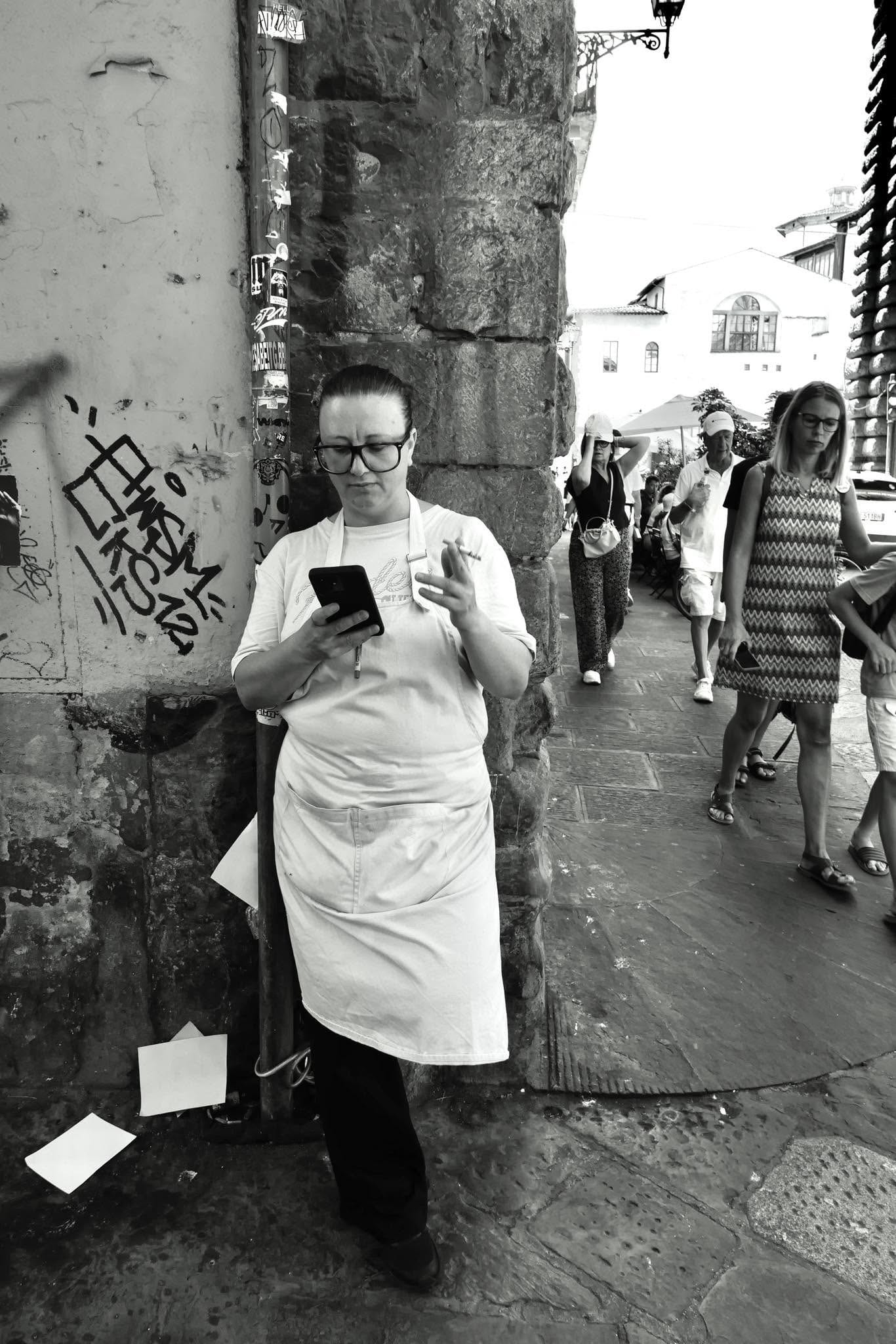 Eine Kellnerin in weißer Schürze lehnt in einer Gasse an der Wand und schaut aufs Telefon
