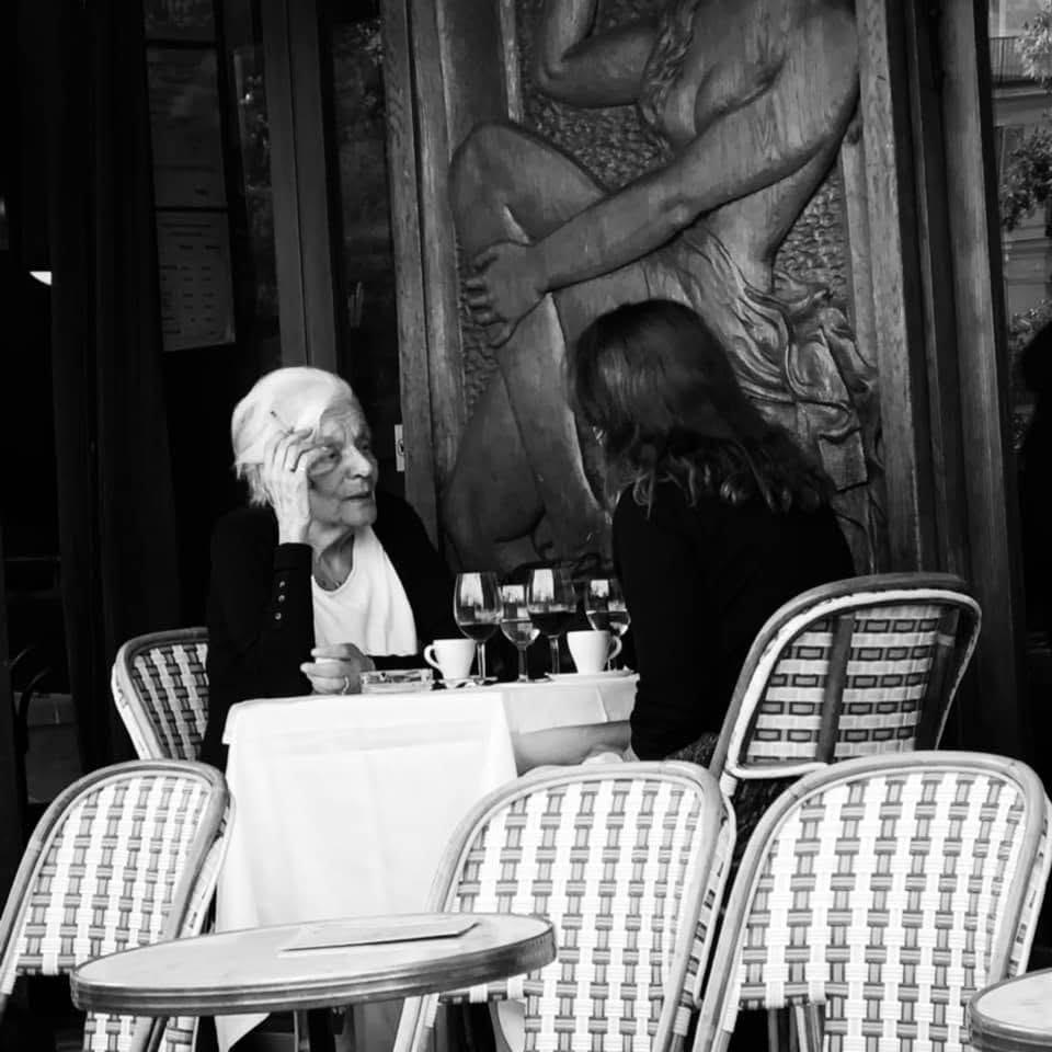 Eine ältere Frau sitzt an einem Cafétisch, die Hand am Kopf, neben ihr ein Relief